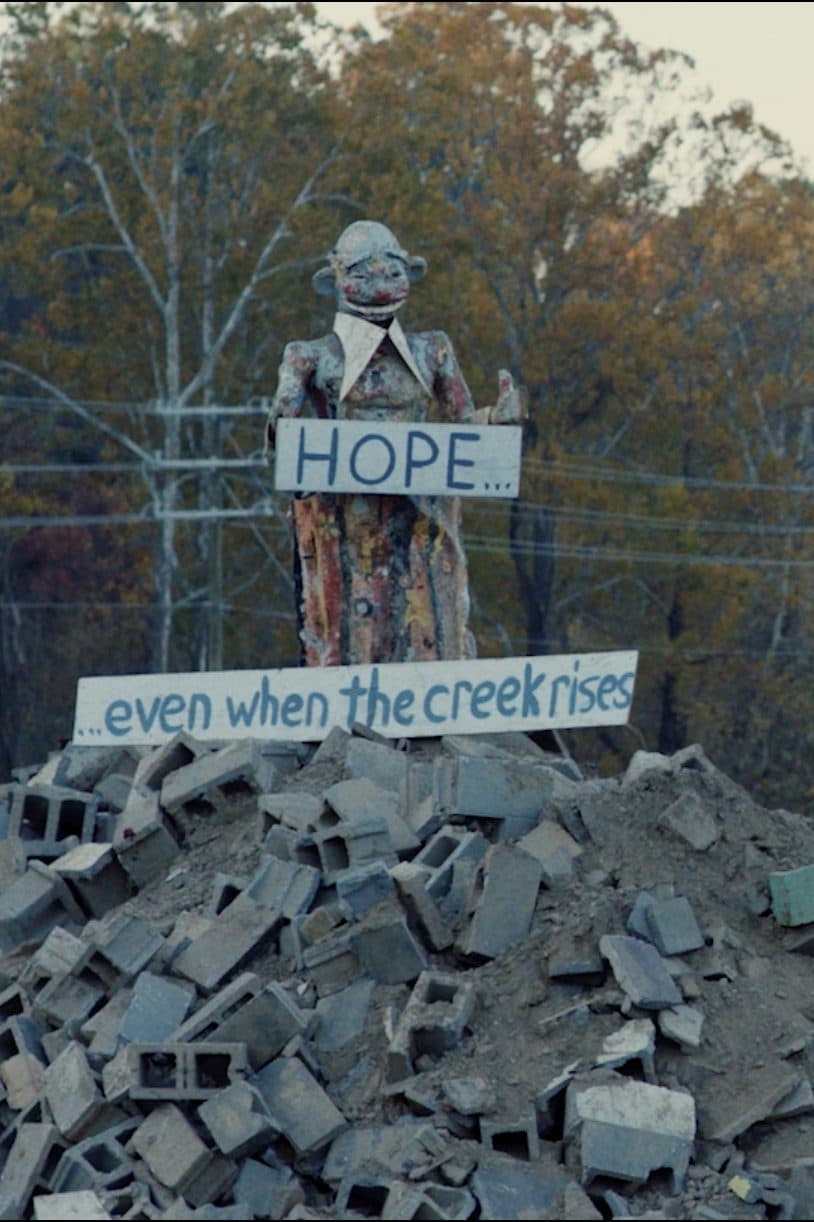 A figure on a pile of rubble holding a sign: “Hope… even when the creek rises.”
