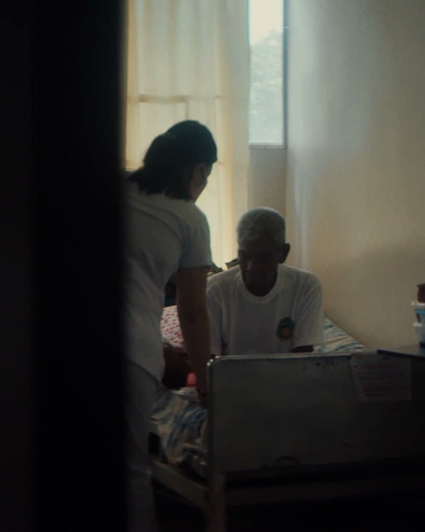 A patient and a nurse in a ward, through a dark doorway.